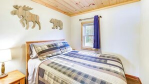 A bedroom with a wooden ceiling, a bed with a plaid bedspread, a nightstand with a lamp, and wall art of a moose and a bear.