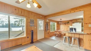 A wooden cabin interior with a large window overlooking a beach, a dining area, and a kitchen with a refrigerator.