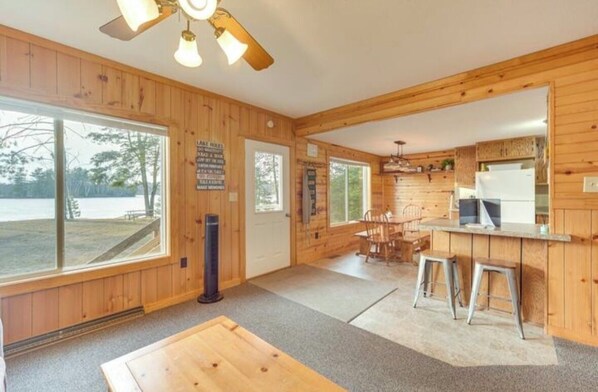 A wooden cabin interior with a large window overlooking a beach, a dining area, and a kitchen with a refrigerator.