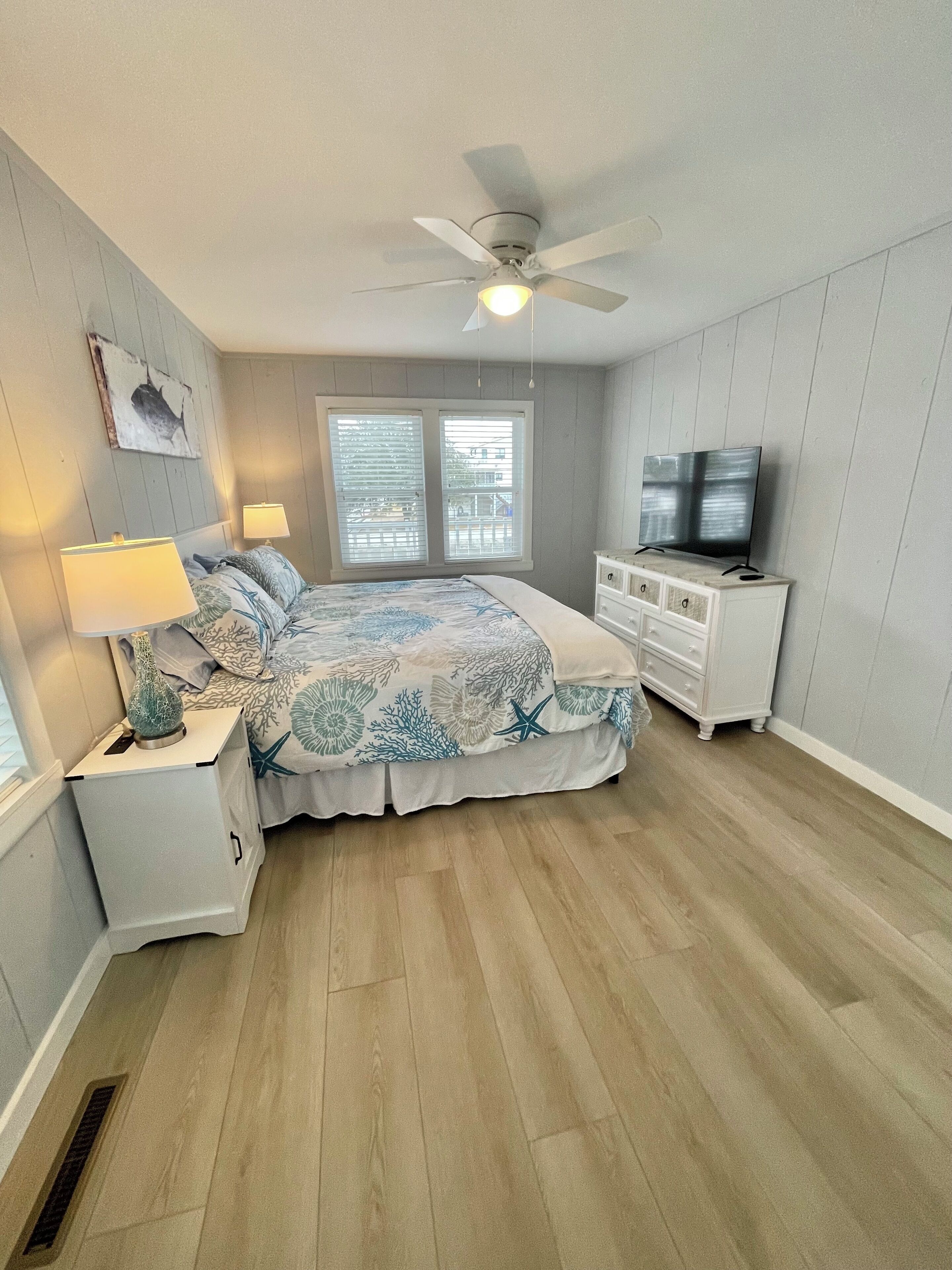 Une chambre à coucher comprenant un lit, des tables de chevet, un téléviseur, un ventilateur de plafond et une fenêtre avec des stores.