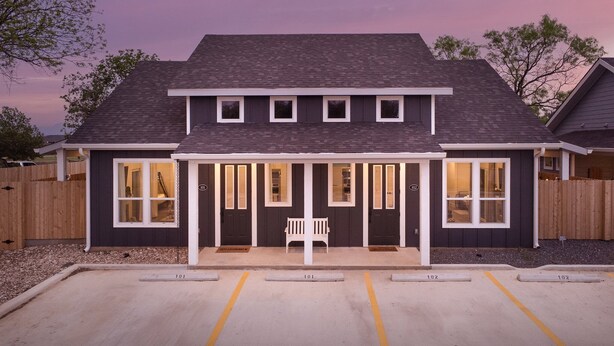 A two-story house with a covered porch, numbered doors, and a parking area in front.
