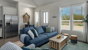 A modern living room with a blue sofa, a wooden coffee table, and a kitchen area with a stainless steel refrigerator.