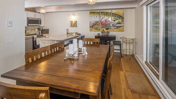 A spacious dining area with a wooden table and chairs, a kitchen in the background, and a large window with a view.