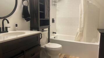 Un baño con lavamanos blanco, mueble de madera oscura, inodoro y bañera blanca con cortina de ducha.