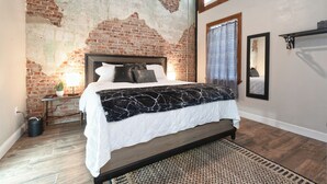Ein Schlafzimmer mit Backsteinwand, einem Bett mit weiß-schwarz gemustertem Überwurf, einem kleinen Nachttisch mit Lampe und einem großen Spiegel an der Wand.