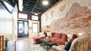Ein modernes Wohnzimmer mit einem roten Sofa, einem gemusterten Sessel und einem Couchtisch. Eine Backsteinwand und ein großes Fenster sind zu sehen.