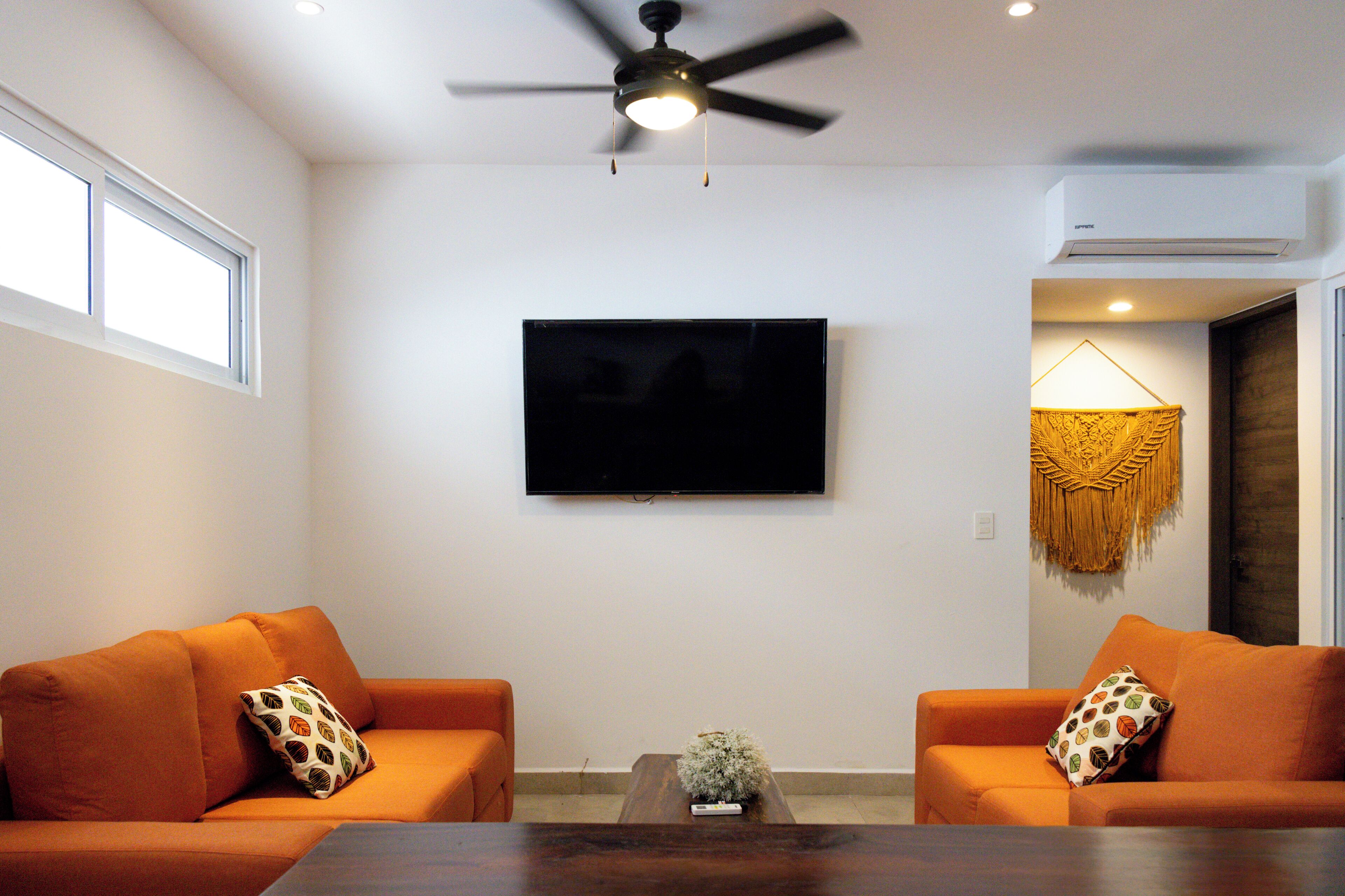 Una sala de estar con dos sofás naranja, un televisor de pantalla plana, un ventilador de techo y un equipo de aire acondicionado.
