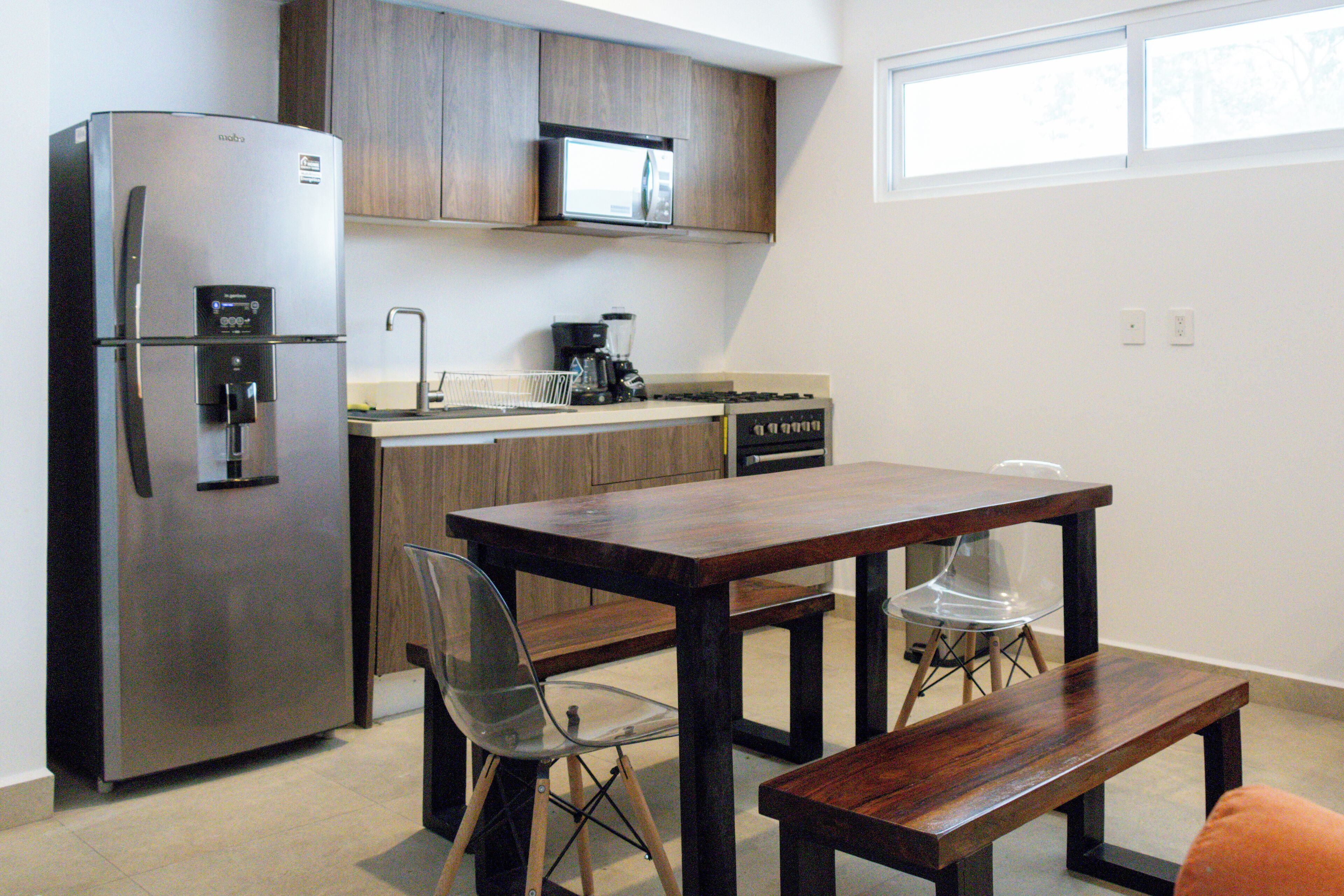 A compact kitchen with a stainless steel refrigerator, a microwave, and a coffee maker on the counter. There is a wooden dining table with two chairs and a bench.