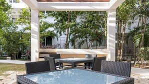 An outdoor patio area with a glass-top table and wicker chairs, surrounded by lush greenery and a white building in the background. - Modern Apt Tulum with 2 BDR Gym & Pool (Tulum)
