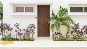 A modern building with a brown door, white walls, and a small window above the door. There are plants and a palm tree in front of the building. - Modern Apt Tulum with 2 BDR Gym & Pool (Tulum)
