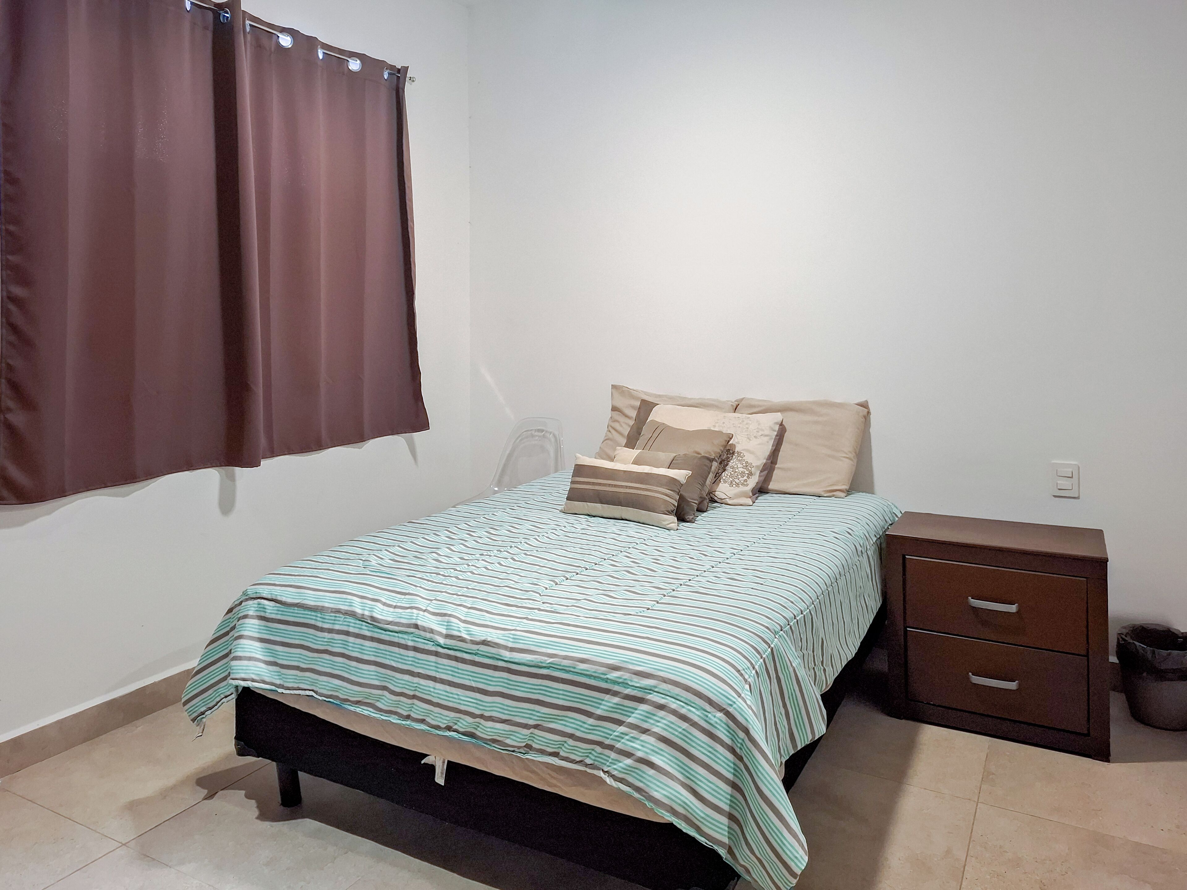 A bedroom with a bed, a nightstand, a chair, and a window with curtains.