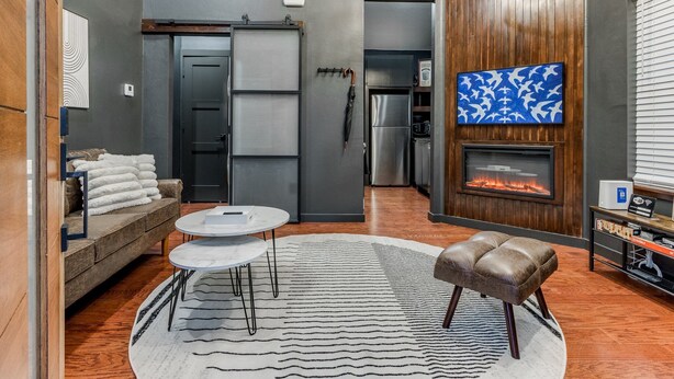A modern living room with a fireplace, a television, and a cozy seating area with a sofa and ottoman. - The Iverson - Urban Core OKC (Oklahoma City)