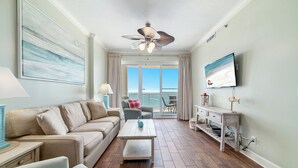 Ein Wohnzimmer mit Sofa, Couchtisch und einem an der Wand montierten Fernseher. Es gibt einen Deckenventilator, ein Fenster mit Vorhängen und einen Blick ins Freie.