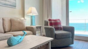 Ein Zimmer mit einem Holztisch, einer blauen Walfigur, einem beige-farbenen Sofa, einem grauen Sessel mit kariertem Kissen, einer Lampe und Meerblick durch ein großes Fenster.