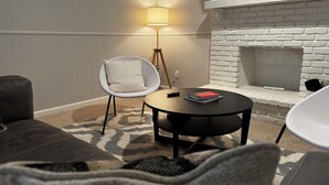 Een gezellige woonkamer met een witte bakstenen open haard, een ronde salontafel, een witte stoel en een vloerkleed met dessin.