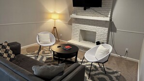 Een woonkamer met een wit bakstenen haardvuur, een flatscreen televisie erboven, een bank met decoratieve kussens, twee witte fauteuils en een vloerlamp.