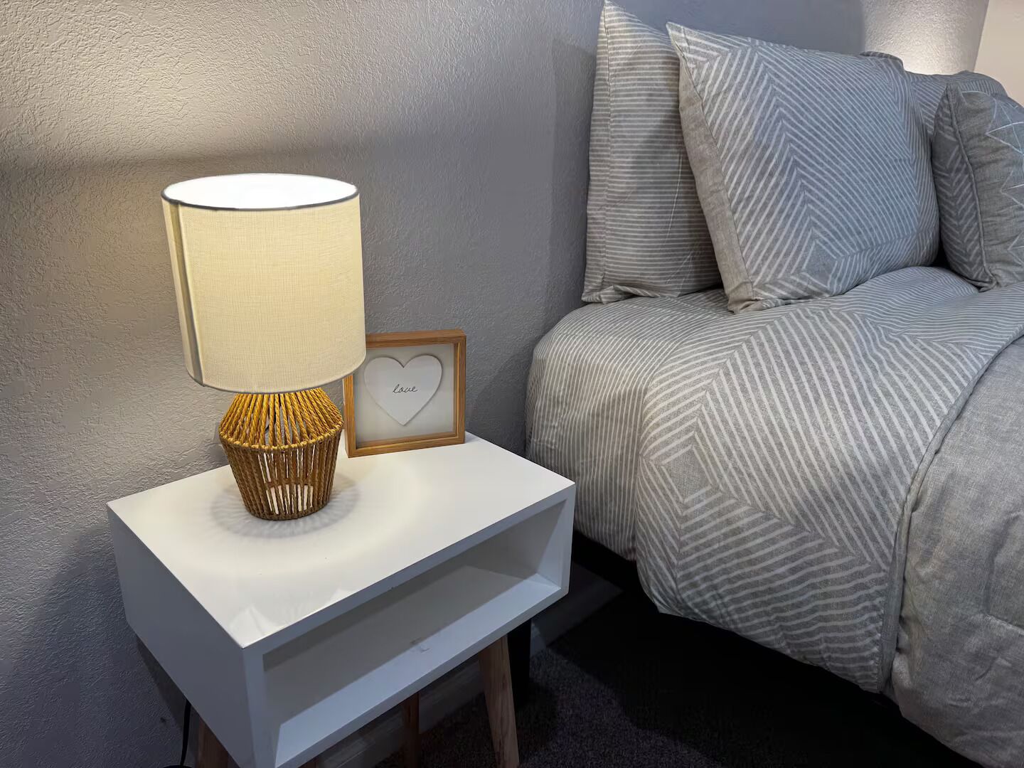 A neatly arranged bedroom with a bedside table, a lamp, a framed picture with a heart, and a bed with striped bedding.