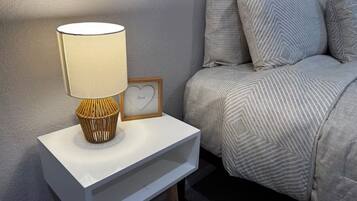 A neatly arranged bedroom with a bedside table, a lamp, a framed picture with a heart, and a bed with striped bedding.