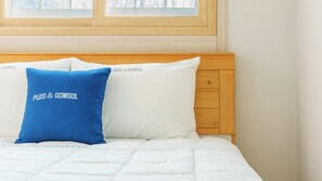 Una cama con colchón blanco y dos almohadas, una azul y otra blanca, en una habitación con cabecera de madera y una ventana.