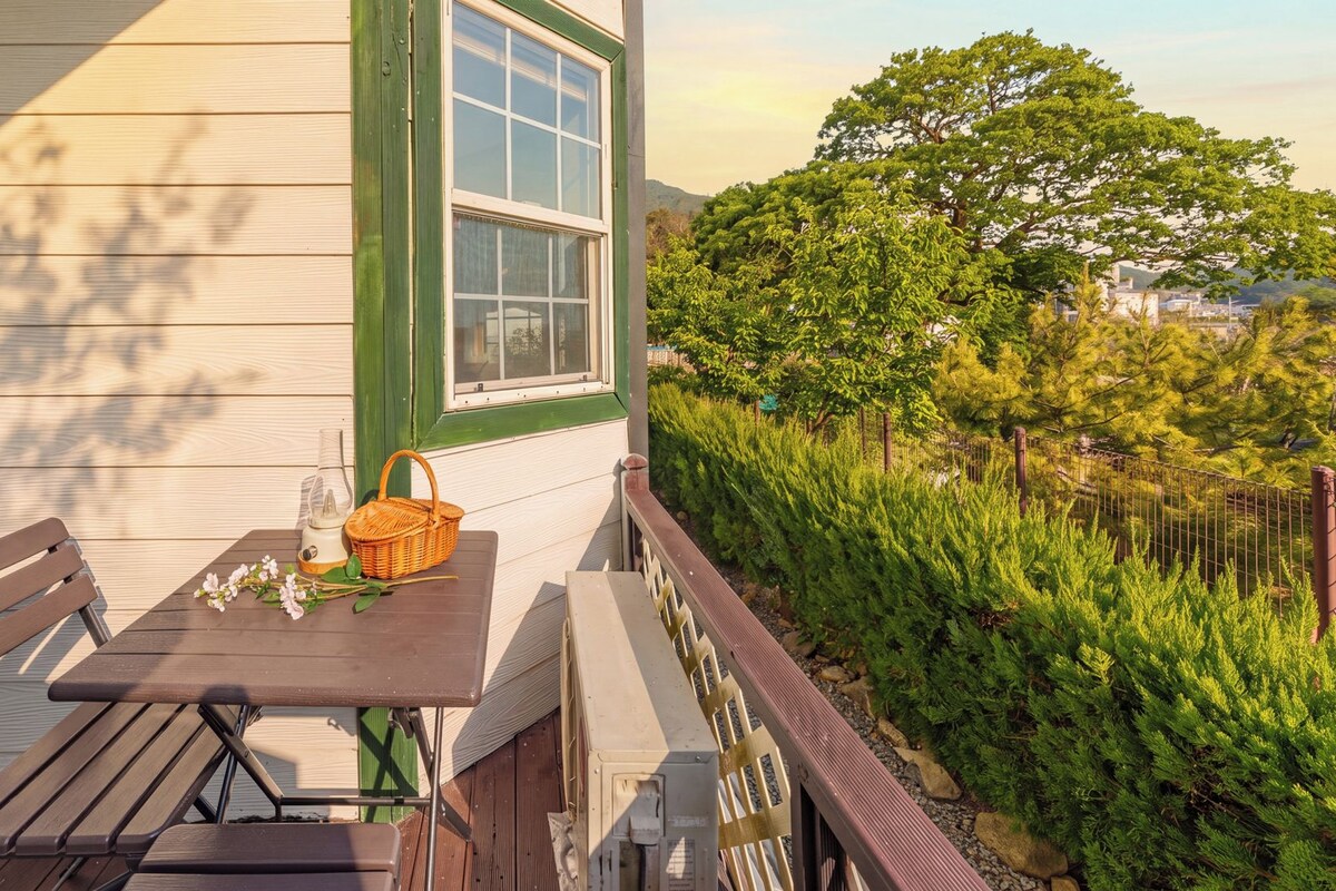 Una terraza de madera con una mesa y bancos, una cesta de mimbre y una ventana con vistas a un área verde exuberante.