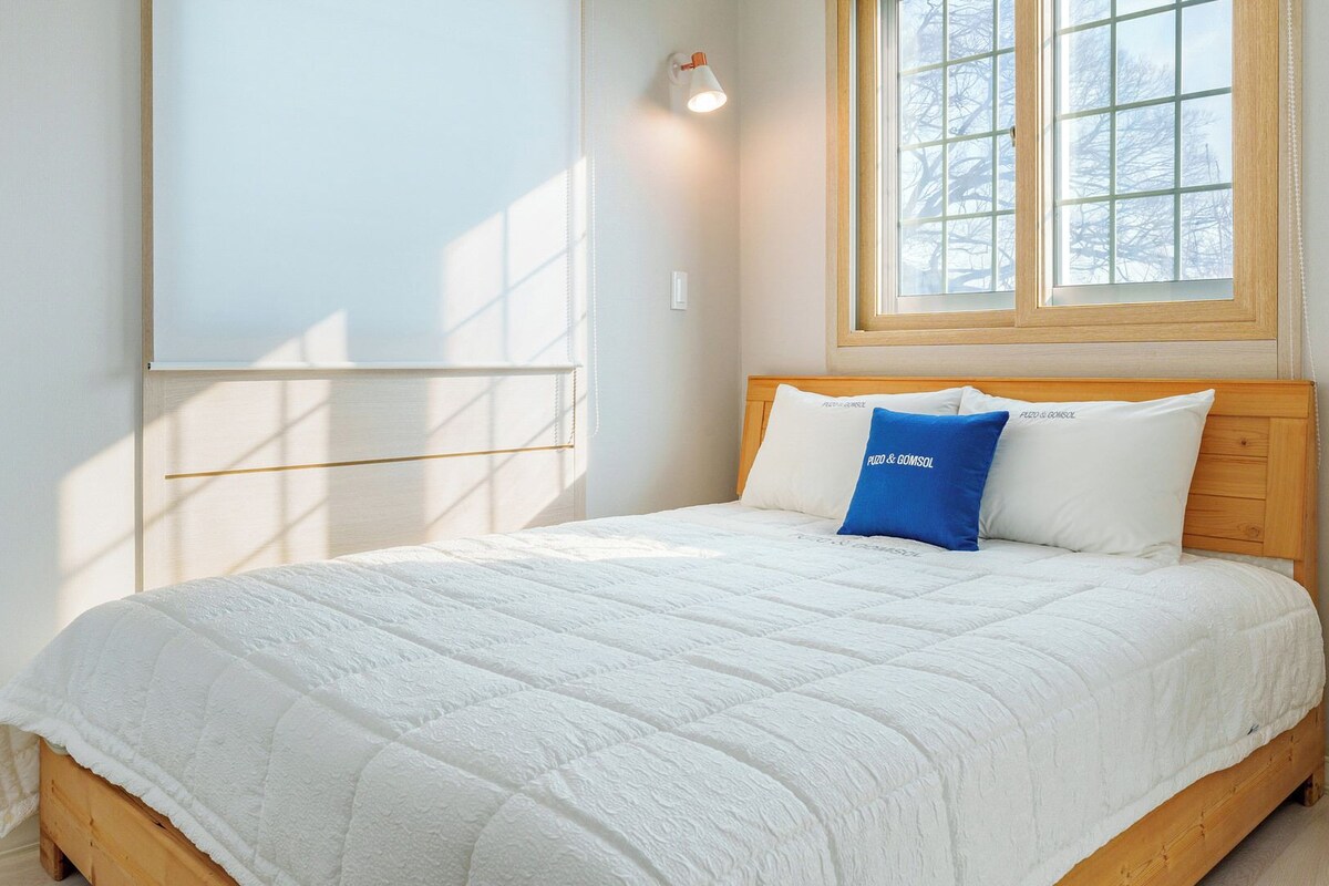 Un dormitorio con cama de madera, ropa de cama blanca, almohada azul y ventana con vistas a árboles.
