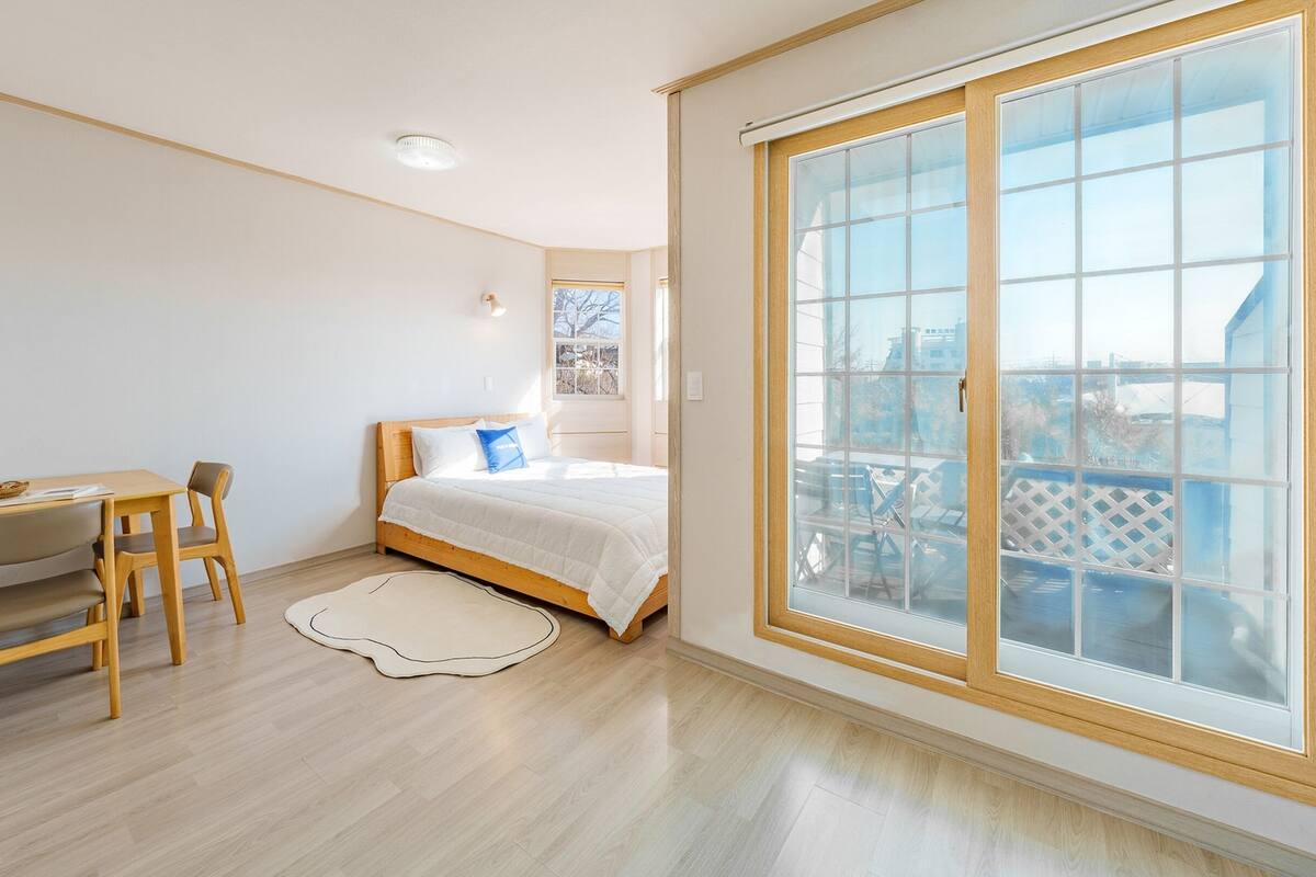 Ein Schlafzimmer mit einer großen Glasschiebetür zu einem Balkon, einem hölzernen Bett, einem Schreibtisch mit Stuhl und einem kleinen Teppich.