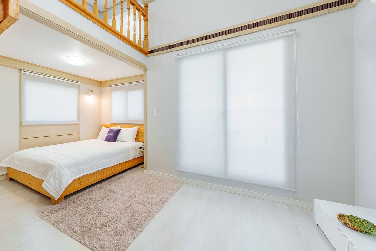 Ein Schlafzimmer mit einem Bett, einem Holz-Kopfteil, einem Fenster mit Jalousien, einer Treppe und einer weißen Wand.