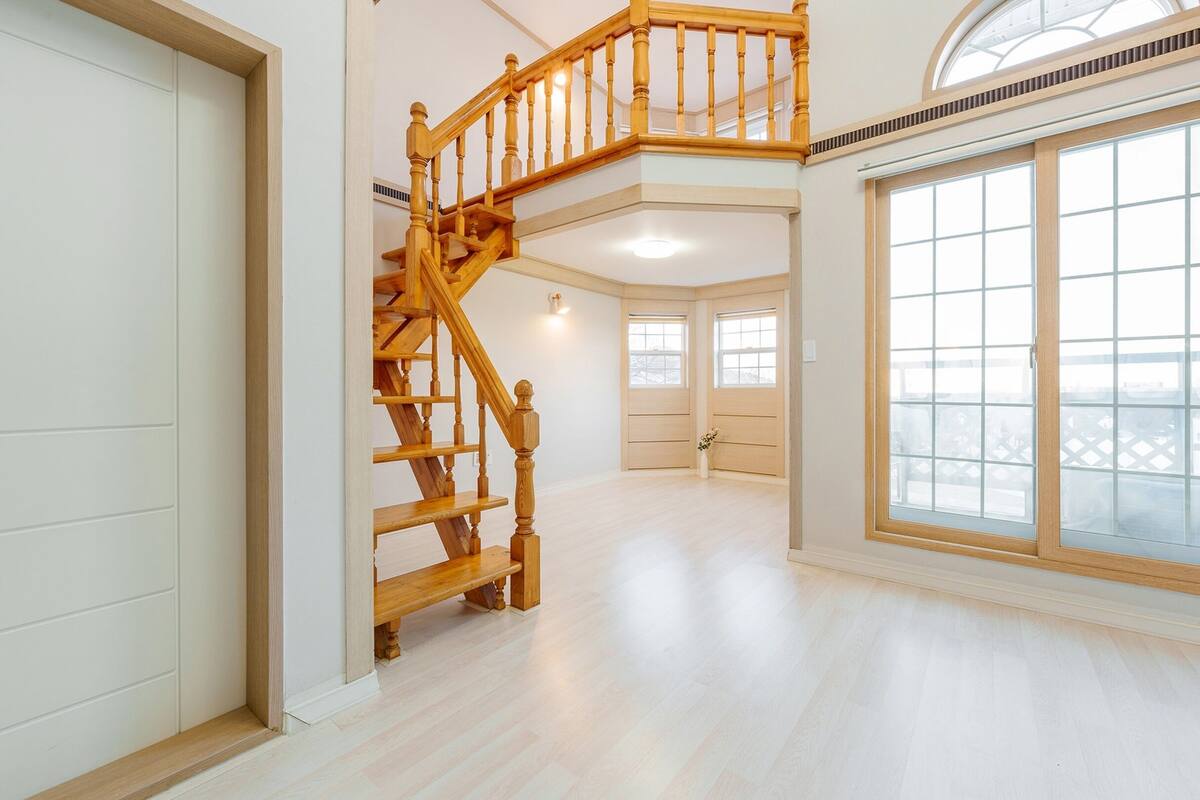 Ein großzügiger Innenraum mit einer Holztreppe zu einer oberen Ebene, großen Fenstern mit Aussicht und einem hellen, freundlichen Boden.
