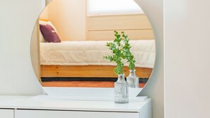 Un miroir reflète un lit avec une tête de lit blanche, une commode blanche ornée de deux vases en verre transparent et une petite plante verte.