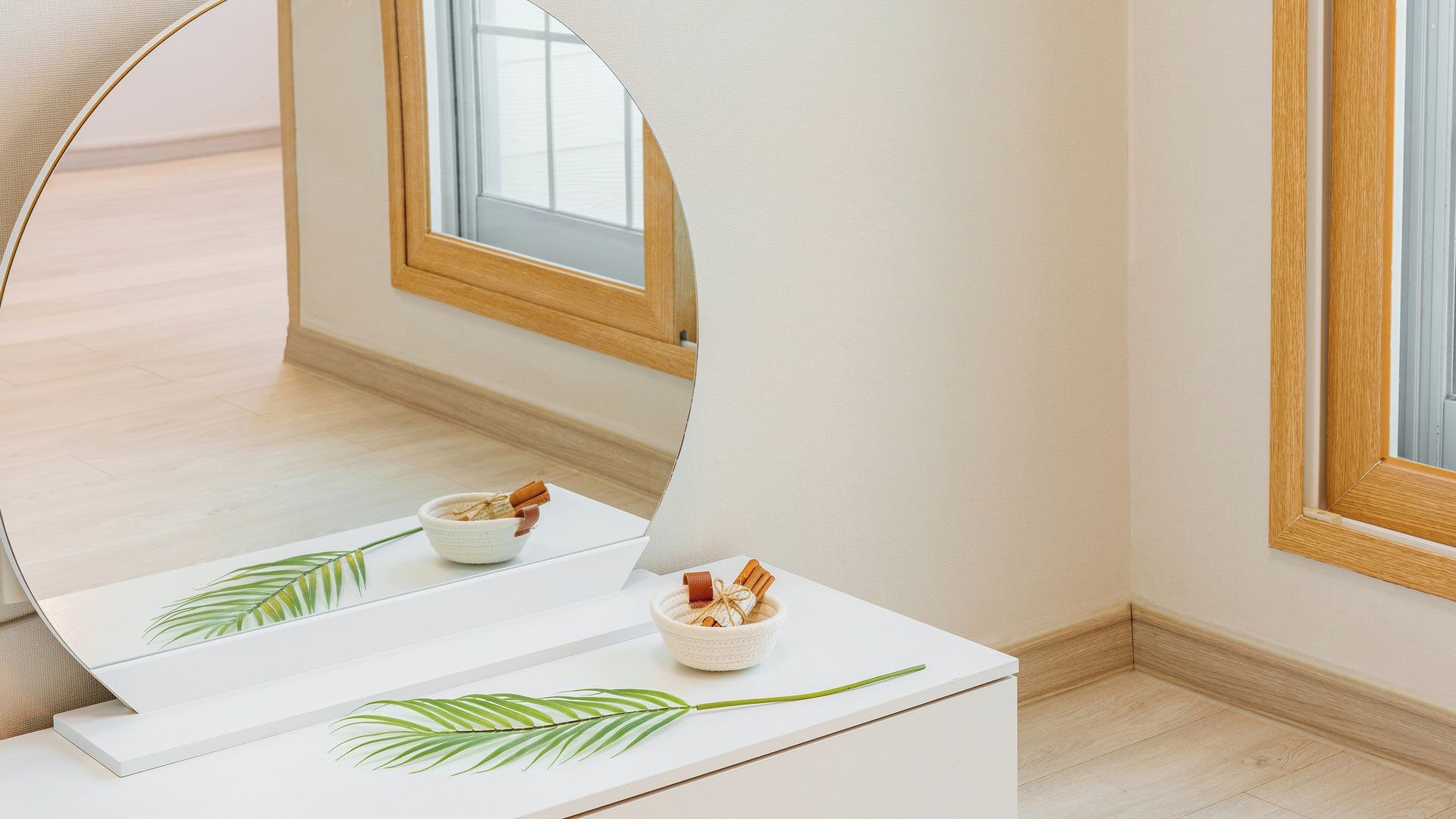 Une pièce avec un grand miroir qui reflète une fenêtre, une table blanche sur laquelle se trouvent un bol et des bâtons de cannelle, et une décoration faite de feuilles vertes.