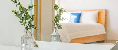 Un dormitorio con una cama, almohadas y dos jarrones con follaje verde sobre una mesita de noche.