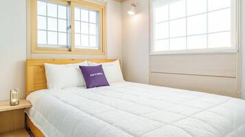 A bedroom with a bed, two windows, a bedside table, and a wall-mounted lamp.