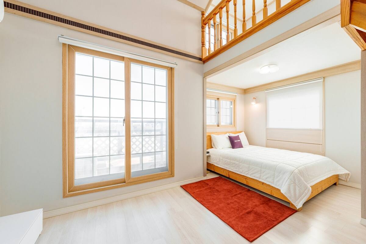 Ein Schlafzimmer mit einer großen Schiebeglaswand, einem Bett mit weißer Bettwäsche, einem roten Teppich und einer Holztreppe.