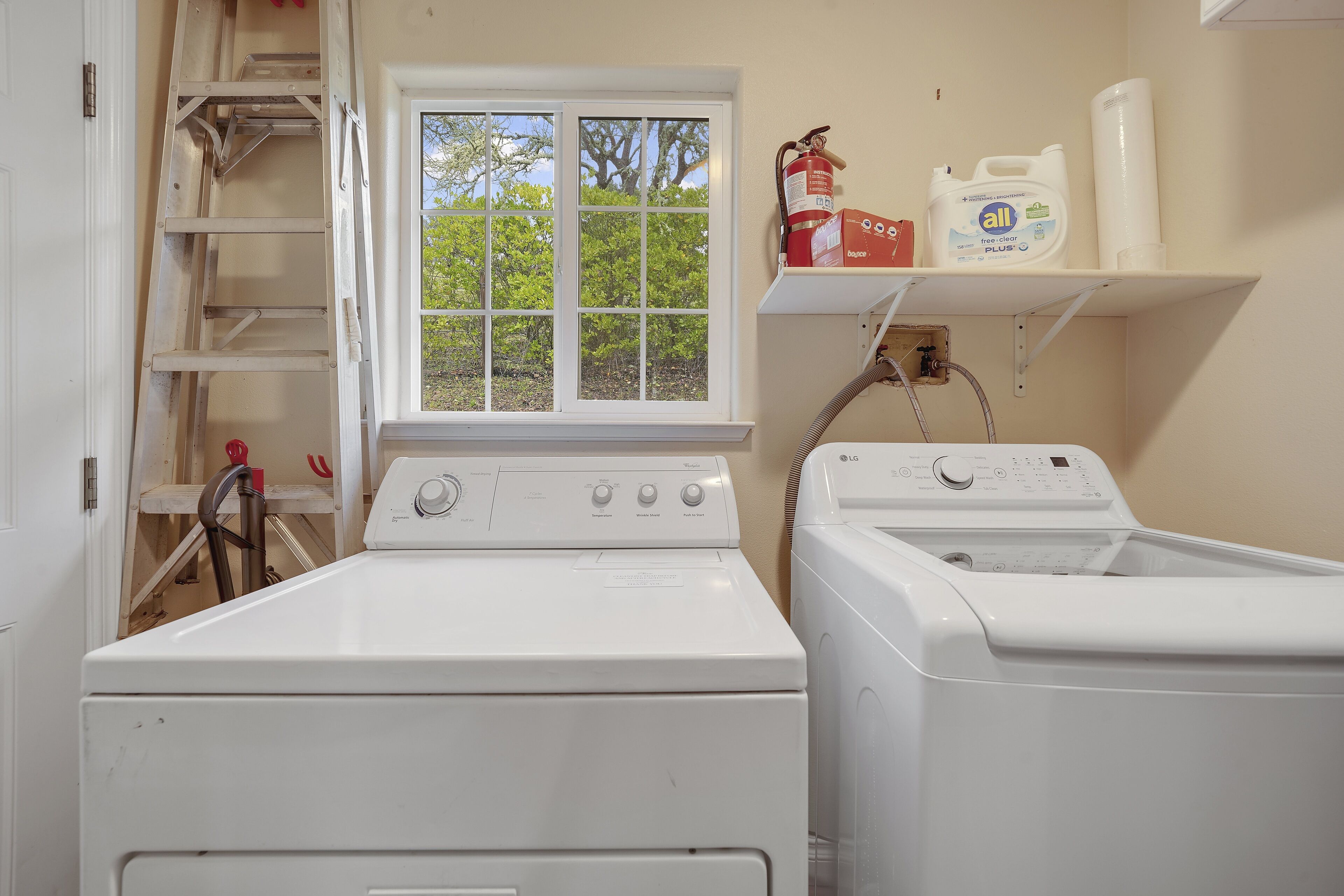 Une buanderie équipée de deux machines à laver et deux sèche-linge, d’une fenêtre et d’une étagère contenant des produits de nettoyage.