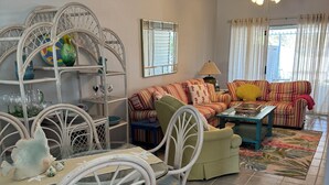 A spacious living room with a dining area, featuring a white wicker dining set, a striped sofa, and a ceiling fan. - Charming retreat across from Beach access 18 and steps to local attractions.  (Panama City Beach)
