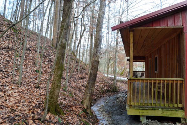 A cabin with a wooden deck and a porch, surrounded by a forested area with a dirt path.