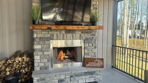 Una chimenea de piedra con un televisor encima, fuego ardiendo y vista a los árboles fuera.