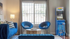 Een woonkamer met een blauwe bank, twee blauwe stoelen, een klein rond tafeltje met een boek, een lamp en een tafelvoetbaltafel.