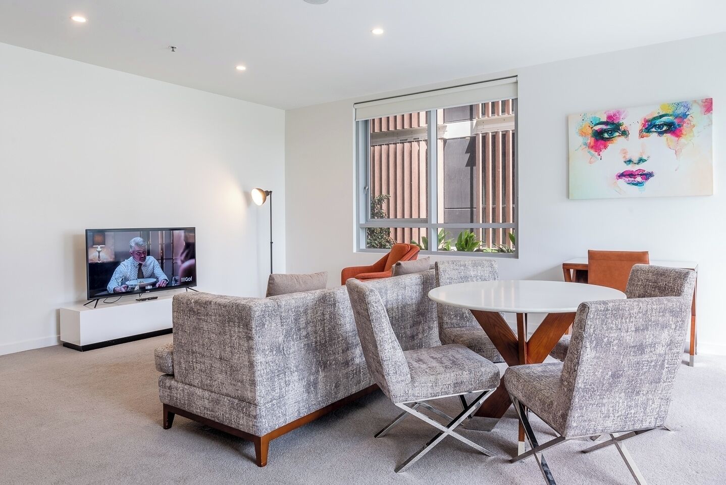 Ein modernes Wohnzimmer mit Flachbildfernseher, grau gepolsterten Sesseln, einem runden Esstisch und einem großen Fenster mit vertikalen Jalousien.