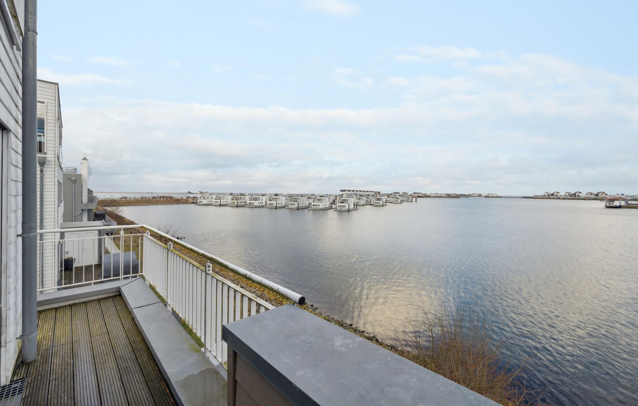 Une propriété située au bord de l’eau, dotée d’un balcon donnant sur une étendue d’eau calme, avec une rangée de bâtiments blancs de l’autre côté et un ciel dégagé.