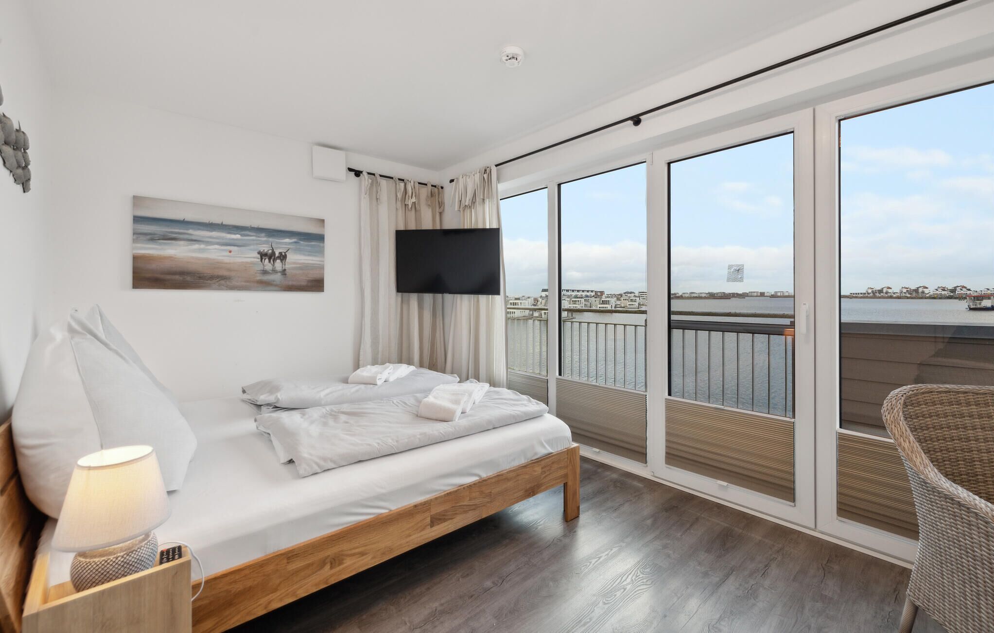 Et moderne hotelværelse med seng, fjernsyn, et maleri og en balkon med udsigt over byen og vandet.