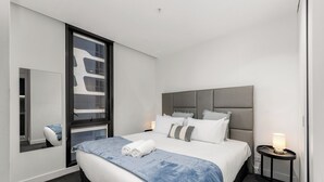 A modern hotel room with a large bed, a bedside table with a lamp, a window with a view, and a mirror on the wall.