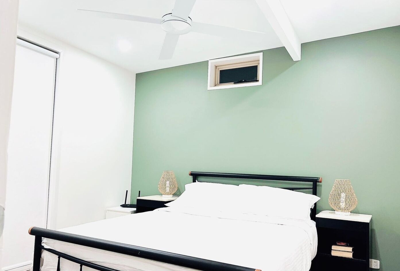 Um quarto com uma cama, duas mesas de cabeceira, um ventilador de teto, uma janela e uma pequena clarabóia.