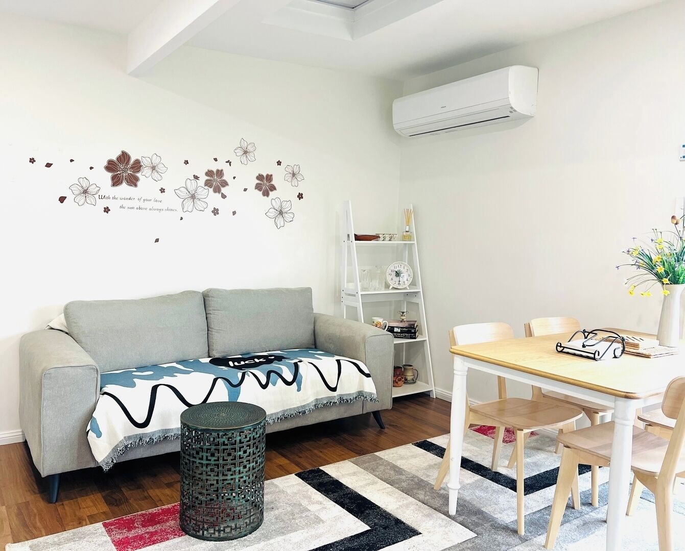 A living room with a sofa, a coffee table, a dining table with chairs, a bookshelf, and an air conditioning unit.