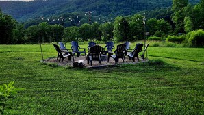An outdoor seating area with chairs and tables, surrounded by a grassy field and a forested hill in the background.