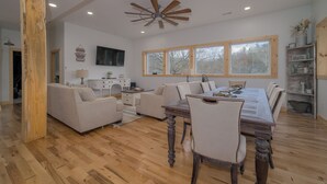 A spacious living room with a wooden floor, a dining table with chairs, a sofa, a TV mounted on the wall, and large windows.