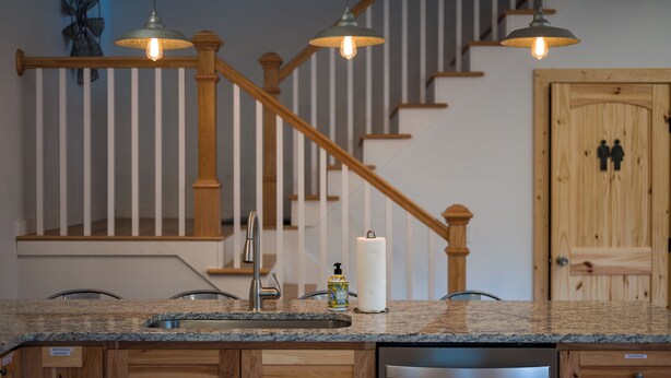 A kitchen with granite countertops, wooden cabinets, a stainless steel dishwasher, and a wooden staircase with a hanging pendant light.