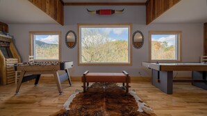 A room with a wooden floor, a large window with a view of trees, a foosball table, and a pool table.