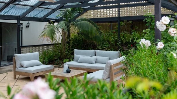 A covered patio area with a wooden sofa set, a coffee table, and lush greenery.