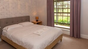 A bedroom with a wooden bed, white linens, a nightstand with a lamp, and a window with curtains.